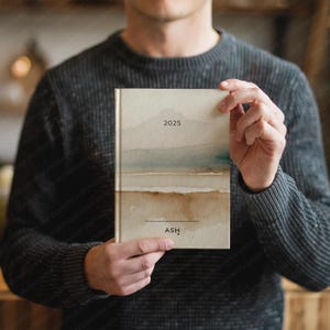 May include: A hardcover journal with a watercolor landscape design in shades of beige, brown, and blue. The year "2025" is printed at the top, and the name "ASH" is printed at the bottom. The journal is held by a person wearing a gray sweater.