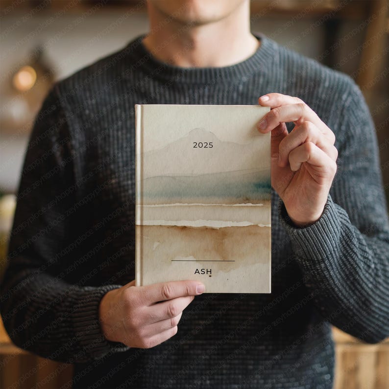 May include: A hardcover journal with a watercolor landscape design in shades of beige, brown, and blue. The year "2025" is printed at the top, and the name "ASH" is printed at the bottom. The journal is held by a person wearing a gray sweater.