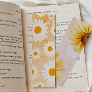 K&ouml;nnte beinhalten: Zwei Lesezeichen mit Aquarell-Blumenmotiven. Ein Lesezeichen zeigt wei&szlig;e G&auml;nsebl&uuml;mchen mit gelben Zentren auf einem Pfirsichhintergrund. Das andere Lesezeichen zeigt eine gelbe Sonnenblume mit braunem Zentrum auf einem Pfirsichhintergrund. Beide Lesezeichen haben einen wei&szlig;en Rand.