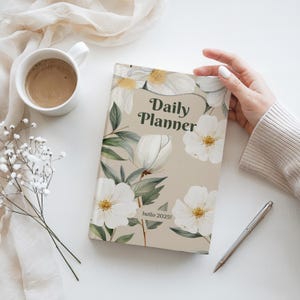 Puede incluir: Una agenda diaria beige con un dise&ntilde;o floral con flores blancas y hojas verdes. El texto "Daily Planner" est&aacute; impreso en verde en la portada. El texto "hello 2025!" est&aacute; impreso en negro en la parte inferior de la portada.