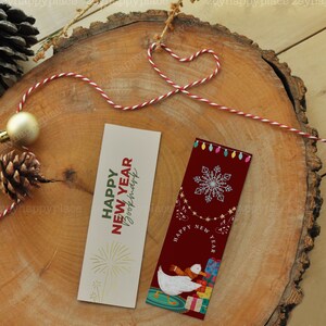 Puede incluir: Dos marcap&aacute;ginas con tem&aacute;tica de A&ntilde;o Nuevo. Un marcap&aacute;ginas es beige con fuegos artificiales dorados y el texto "Happy New Year Bookmark". El otro marcap&aacute;ginas es rojo con un copo de nieve blanco, un ganso blanco con una bufanda y el texto "Happy New Year".