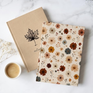 May include: Two notebooks. One has a beige cover with a black maple leaf illustration and the word "autumn" below. The other has a cream cover with a pattern of flowers in brown, yellow, and blue. A cup of coffee sits beside the notebooks.