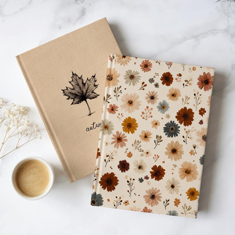 May include: Two notebooks. One has a beige cover with a black maple leaf illustration and the word "autumn" below. The other has a cream cover with a pattern of flowers in brown, yellow, and blue. A cup of coffee sits beside the notebooks.