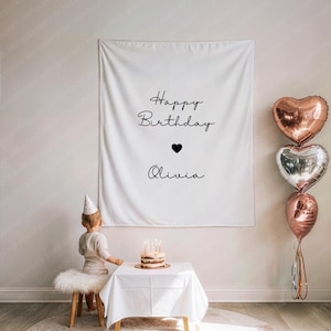 May include: A white fabric banner with the words "Happy Birthday Olivia" in black script, a black heart, and three heart-shaped balloons. A small cake with lit candles sits on a table. A child wearing a party hat is in the foreground.
