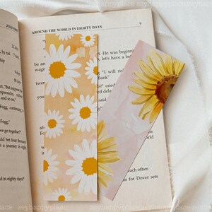 K&ouml;nnte beinhalten: Zwei Lesezeichen mit Blumenmustern. Das erste Lesezeichen zeigt ein wei&szlig;es G&auml;nsebl&uuml;mchenmuster auf einem gelben und orangen Hintergrund. Das zweite Lesezeichen zeigt eine gelbe Sonnenblume auf einem rosa Hintergrund. Beide Lesezeichen haben einen Aquarelleffekt.