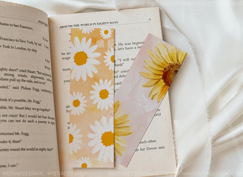 K&ouml;nnte beinhalten: Zwei Lesezeichen mit Blumenmustern. Das erste Lesezeichen zeigt ein wei&szlig;es G&auml;nsebl&uuml;mchenmuster auf einem gelben und orangen Hintergrund. Das zweite Lesezeichen zeigt eine gelbe Sonnenblume auf einem rosa Hintergrund. Beide Lesezeichen haben einen Aquarelleffekt.