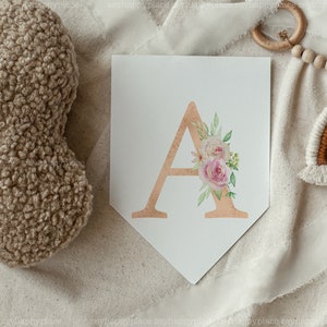 May include: A white pennant banner with a gold letter A decorated with pink and white watercolour flowers. The banner is on a white fabric background.
