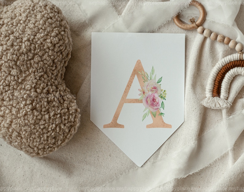 May include: A white pennant banner with a gold letter A decorated with pink and white watercolour flowers. The banner is on a white fabric background.