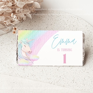 May include: A white card with a rainbow and unicorn design. The card says "Emma is turning 1" in blue and pink text.