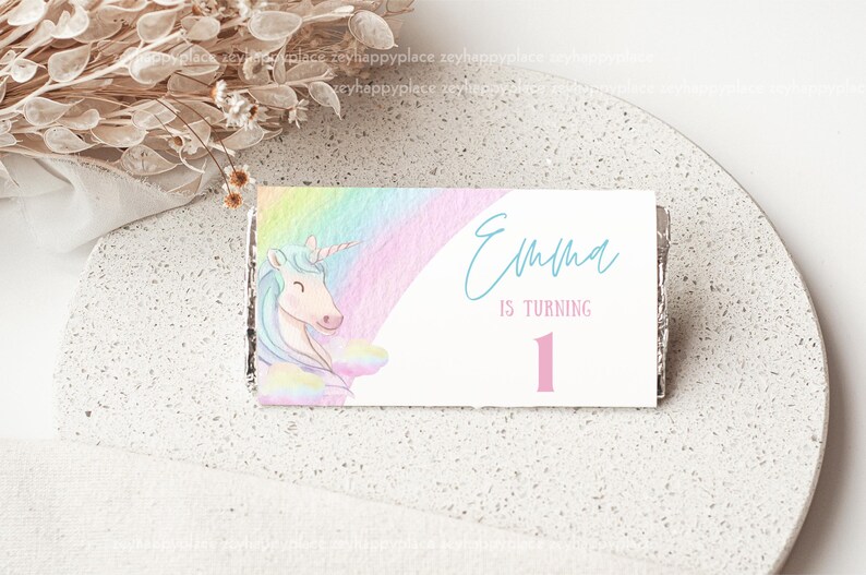 May include: A white card with a rainbow and unicorn design. The card says "Emma is turning 1" in blue and pink text.
