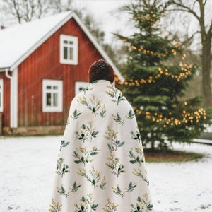 May include: A person wearing a cream-colored blanket with a botanical pattern of yellow and green sprigs. The blanket is draped over the shoulders, with a red house and snow-covered ground in the background. A Christmas tree with lights is also visible.