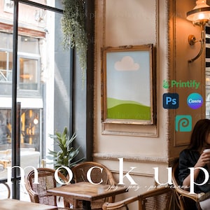 May include: A framed print of a simple landscape with a green hill, a blue sky, and a white cloud. The print is hanging on a wall in a cafe setting with a table and chairs in the foreground.