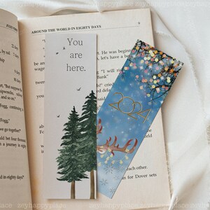 K&ouml;nnte beinhalten: Zwei Lesezeichen mit unterschiedlichen Designs. Das erste Lesezeichen ist wei&szlig; mit einer Aquarell-Illustration von zwei Kiefern und dem Text "You are here." Das zweite Lesezeichen ist blau mit einer goldenen Rentier-Silhouette, goldenem Glitzer und dem Text "2024".