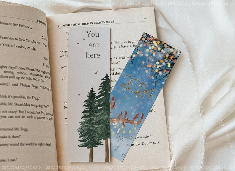 K&ouml;nnte beinhalten: Zwei Lesezeichen mit unterschiedlichen Designs. Das erste Lesezeichen ist wei&szlig; mit einer Aquarell-Illustration von zwei Kiefern und dem Text "You are here." Das zweite Lesezeichen ist blau mit einer goldenen Rentier-Silhouette, goldenem Glitzer und dem Text "2024".