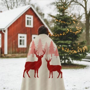 May include: A cream-colored blanket featuring a red deer and pink tree design. The blanket is draped over a person standing in a snowy landscape with a red house and Christmas tree in the background.