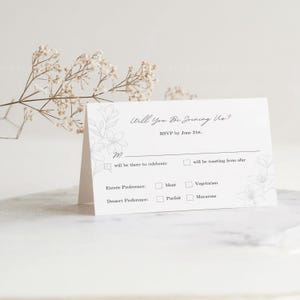 May include: A white RSVP card with a floral design and the text "Will You Be Joining Us?" RSVP by June 21st.  The card has checkboxes for guests to indicate their attendance and meal preferences.  The options include "will be there to celebrate", "will be toasting from afar", "Meat", "Vegetarian", "Parfait", and "Macarons".