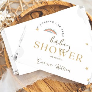 May include: A white card with gold lettering announcing a baby shower for Emma Watson. The card features a rainbow and stars.