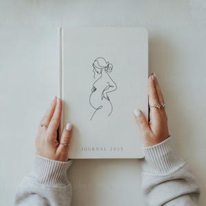 May include: A white journal with a line drawing of a pregnant woman on the cover. The journal has the text "JOURNAL 2025" printed on the bottom.
