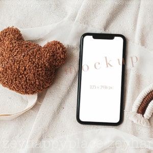 May include: A black smartphone mockup with a white screen on a beige fabric background. The phone is surrounded by a brown teddy bear head, a white and brown rainbow, and a white ribbon.