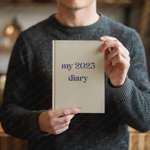 May include: A beige diary with the text "my 2025 diary" in dark blue. The diary is held by a person wearing a dark gray sweater. The year "2025" is also printed at the bottom of the diary.