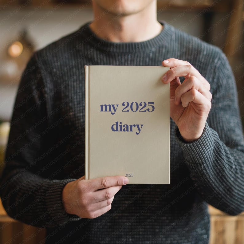 May include: A beige diary with the text "my 2025 diary" in dark blue. The diary is held by a person wearing a dark gray sweater. The year "2025" is also printed at the bottom of the diary.