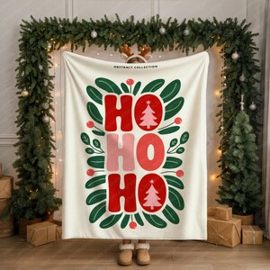 May include: A cream-colored blanket with the words "HO HO HO" in red and pink, with a Christmas tree design. The blanket is surrounded by green leaves and red berries, with the text "ABSTRACT COLLECTION" at the top.