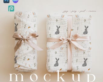 May include: Two white muslin baby blankets with a bunny rabbit and leaf pattern. The blankets are tied with a beige ribbon and are on a light beige background. The text "Mockup" is printed below the blankets.