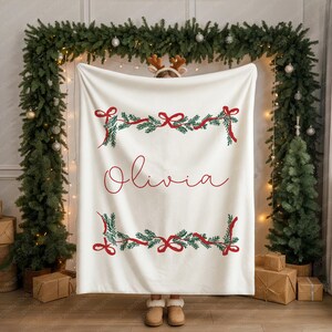 May include: A cream-colored blanket with red and green Christmas garland designs and the name "Olivia" in red script. The blanket is held up in front of a Christmas-themed backdrop with a garland and small Christmas trees.