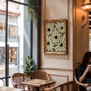 K&ouml;nnte beinhalten: Ein gerahmter Druck eines stilisierten Blumendesigns mit wei&szlig;en Bl&uuml;ten auf gr&uuml;nem Hintergrund. Der Druck h&auml;ngt an einer Wand in einem Caf&eacute; mit Holzst&uuml;hlen und einem Tisch.
