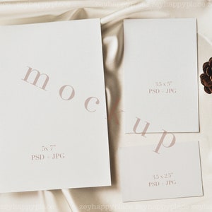 May include: Three blank white cards on a cream-colored fabric background. The cards are different sizes: 5x7 inches, 3.5x5 inches, and 3.5x2.5 inches. The text "mockup" is written in a light pink font across the cards. A brown pine cone is on the fabric to the right of the cards.