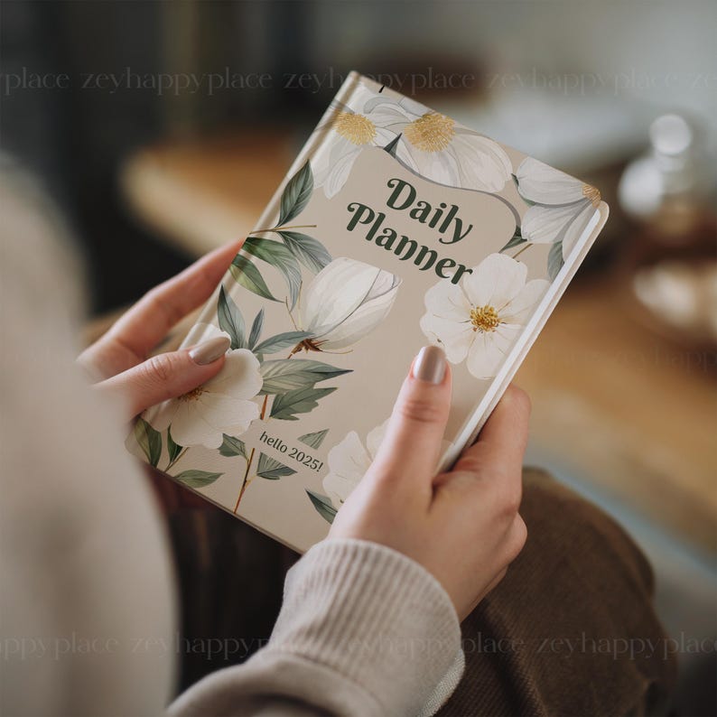 May include: A beige daily planner with a floral design featuring white flowers and green leaves. The text "Daily Planner" is written in a cursive font on the front cover. The text "hello 2025!" is written in a smaller font at the bottom of the cover.