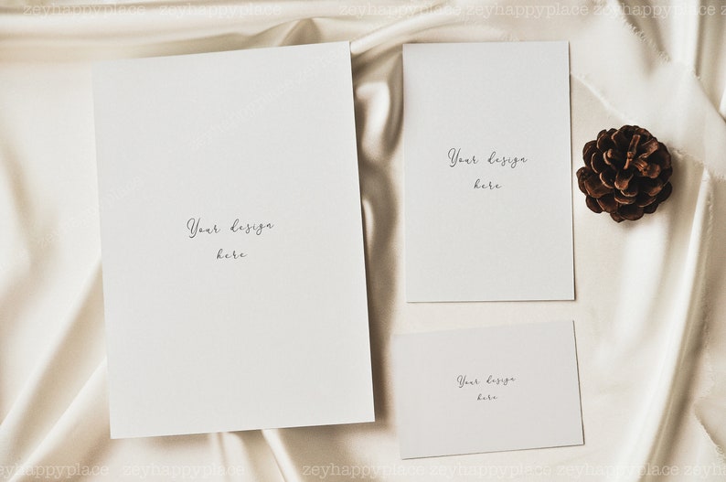 May include: Three blank cards with the text "Your design here" on a white satin background. A brown pine cone is on the right side of the image.