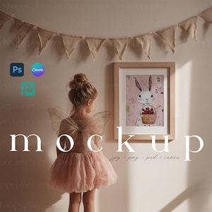 Puede incluir: Una obra de arte enmarcada con un conejo blanco sosteniendo una cesta de cerezas cuelga de una pared. Una persona joven con un tutú rosa y alas de hada está de pie frente a la obra de arte. La palabra "mockup" se muestra en letras grandes.