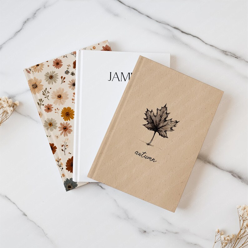 May include: Three notebooks with different covers. One has a floral pattern in beige, brown, and yellow. Another is white with the name "JAM" printed on it. The third is tan with a maple leaf illustration and the word "autumn".