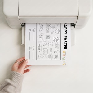 May include: A printer is outputting a black and white Easter-themed coloring page. The page features various Easter-related illustrations, including eggs, bunnies, and flowers, with the words "Happy Easter" printed vertically on the right side.