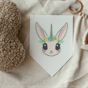 May include: A white bunny with a rainbow horn and a flower crown. The bunny has big, black eyes and a pink nose. The background is a white, textured fabric.
