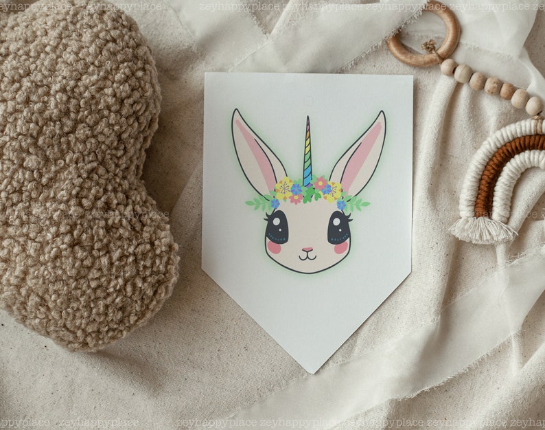 May include: A white bunny with a rainbow horn and a flower crown. The bunny has big, black eyes and a pink nose. The background is a white, textured fabric.