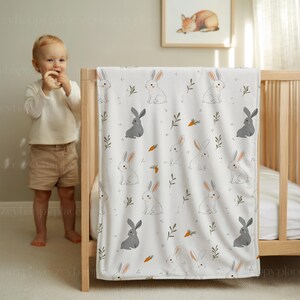 May include: A white baby blanket featuring a pattern of grey and white rabbits, carrots, and green sprigs. The blanket is draped over a wooden cot. A framed picture of a fox is on the wall.