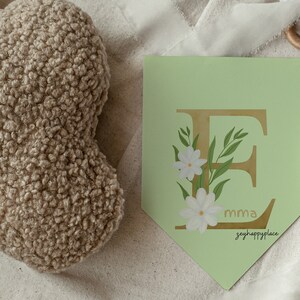 May include: A light green banner with a gold letter E decorated with white flowers and green leaves. The banner has the text "mma" and "zeyhappylace" printed on it.