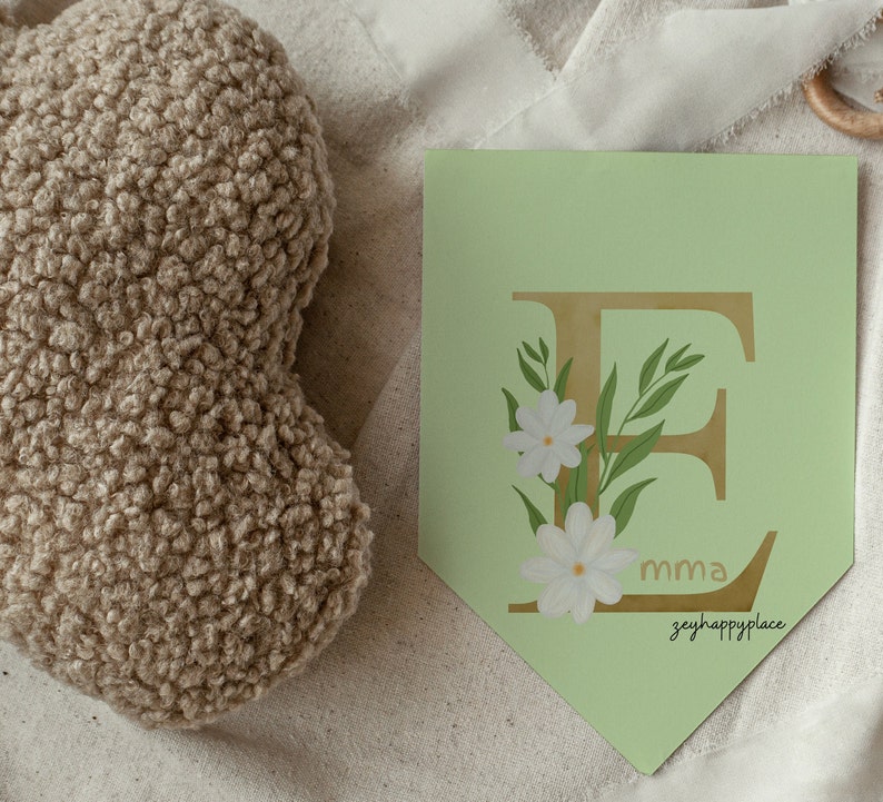 May include: A light green banner with a gold letter E decorated with white flowers and green leaves. The banner has the text "mma" and "zeyhappylace" printed on it.