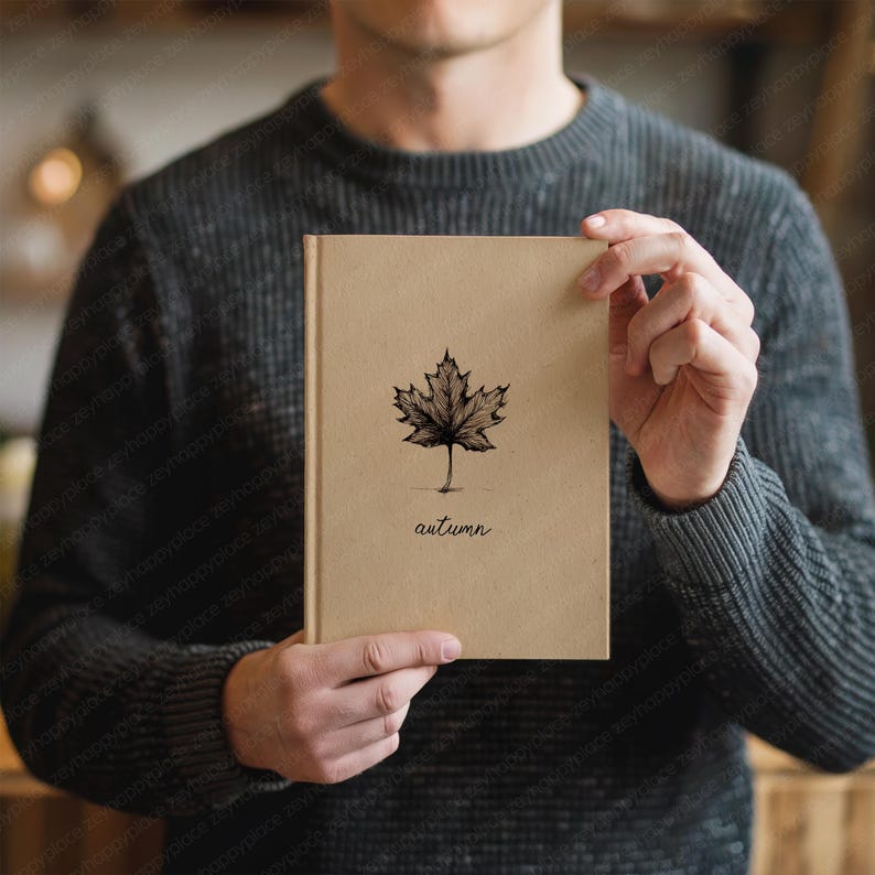 May include: A tan-colored notebook with a black maple leaf illustration and the word "autumn" printed below it. The notebook is held by a person wearing a gray sweater.