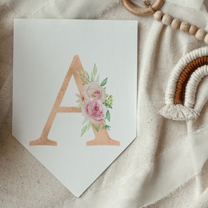 May include: A white pennant banner with a gold letter A decorated with pink and white watercolour flowers. The banner is on a white fabric background.