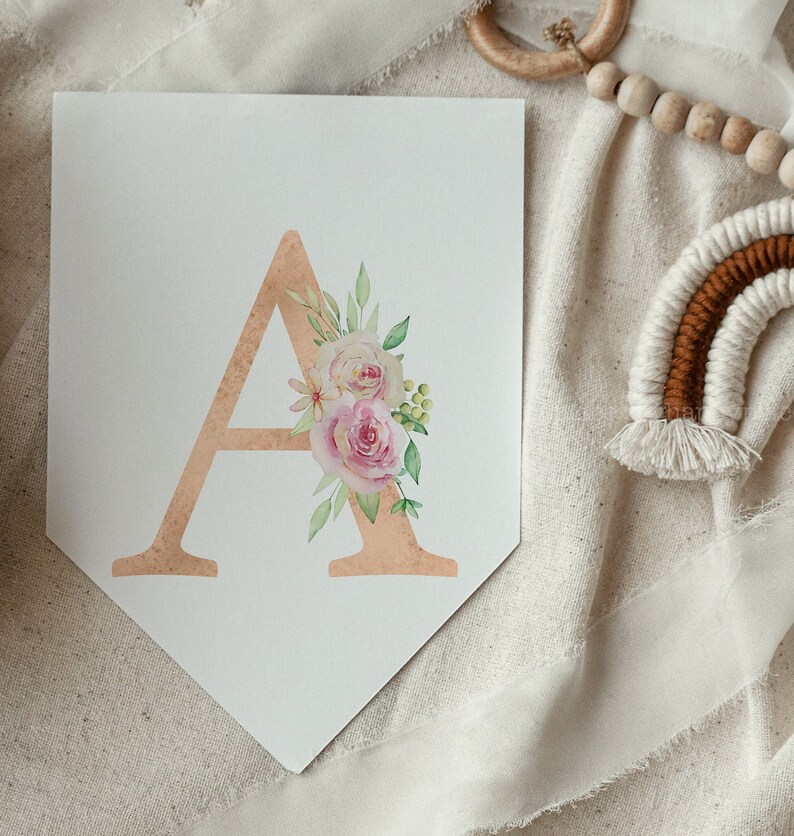 May include: A white pennant banner with a gold letter A decorated with pink and white watercolour flowers. The banner is on a white fabric background.