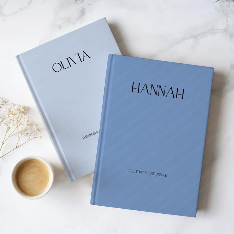May include: Two hardback journals, one pale blue and one periwinkle, with the names "OLIVIA" and "HANNAH" printed on the covers. The pale blue journal has the text "THINGS SHE" and the periwinkle journal has the text "ALL THAT WENT UNSAID". A cup of coffee sits nearby.