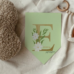 May include: A green and gold letter E with white flowers and green leaves. The letter is on a light green banner with the text "emma" and "greyhappyplace" in gold.