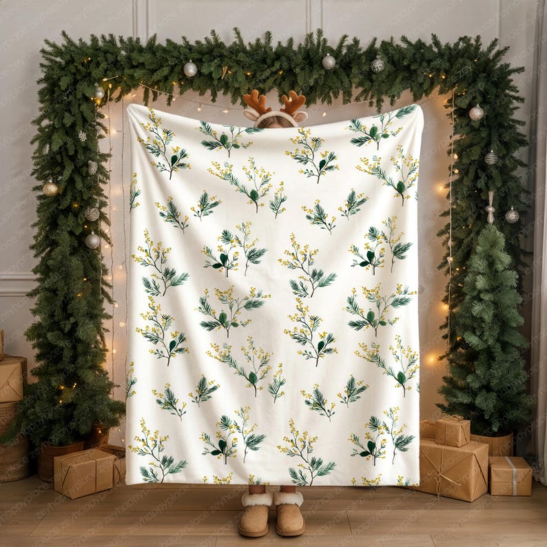 May include: A white blanket with a repeating pattern of green leaves and yellow flowers. The blanket is held up in front of a person. The background includes Christmas decorations, such as a garland and a Christmas tree.