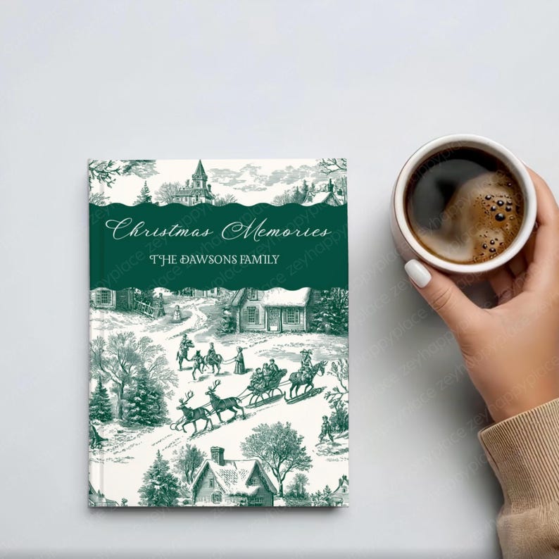 May include: A hardcover Christmas Memories journal with a green and white winter scene illustration. The cover reads "Christmas Memories" and "The Dawsons Family". A cup of coffee is held in the background.