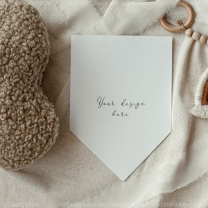 May include: A white card with a pointed bottom edge, lying on a beige fabric surface. The card has the text "Your design here" written on it in black ink. The card is surrounded by a white fabric, a wooden bead garland, and a brown and white macrame rainbow.