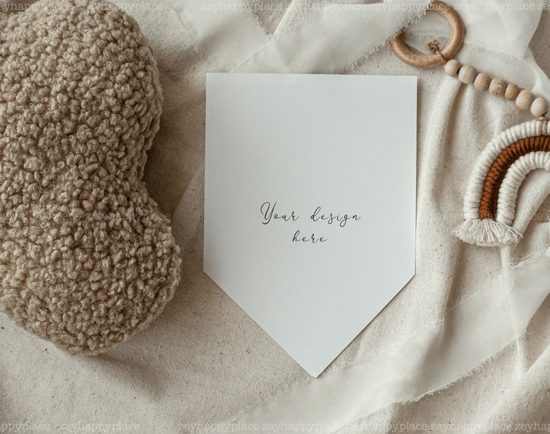 May include: A white card with a pointed bottom edge, lying on a beige fabric surface. The card has the text "Your design here" written on it in black ink. The card is surrounded by a white fabric, a wooden bead garland, and a brown and white macrame rainbow.