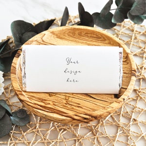 Op de afbeelding: Een witte chocolade reep met een blanco label waarop "Your design here" staat, op een houten dienblad met een geweven onderzetter.